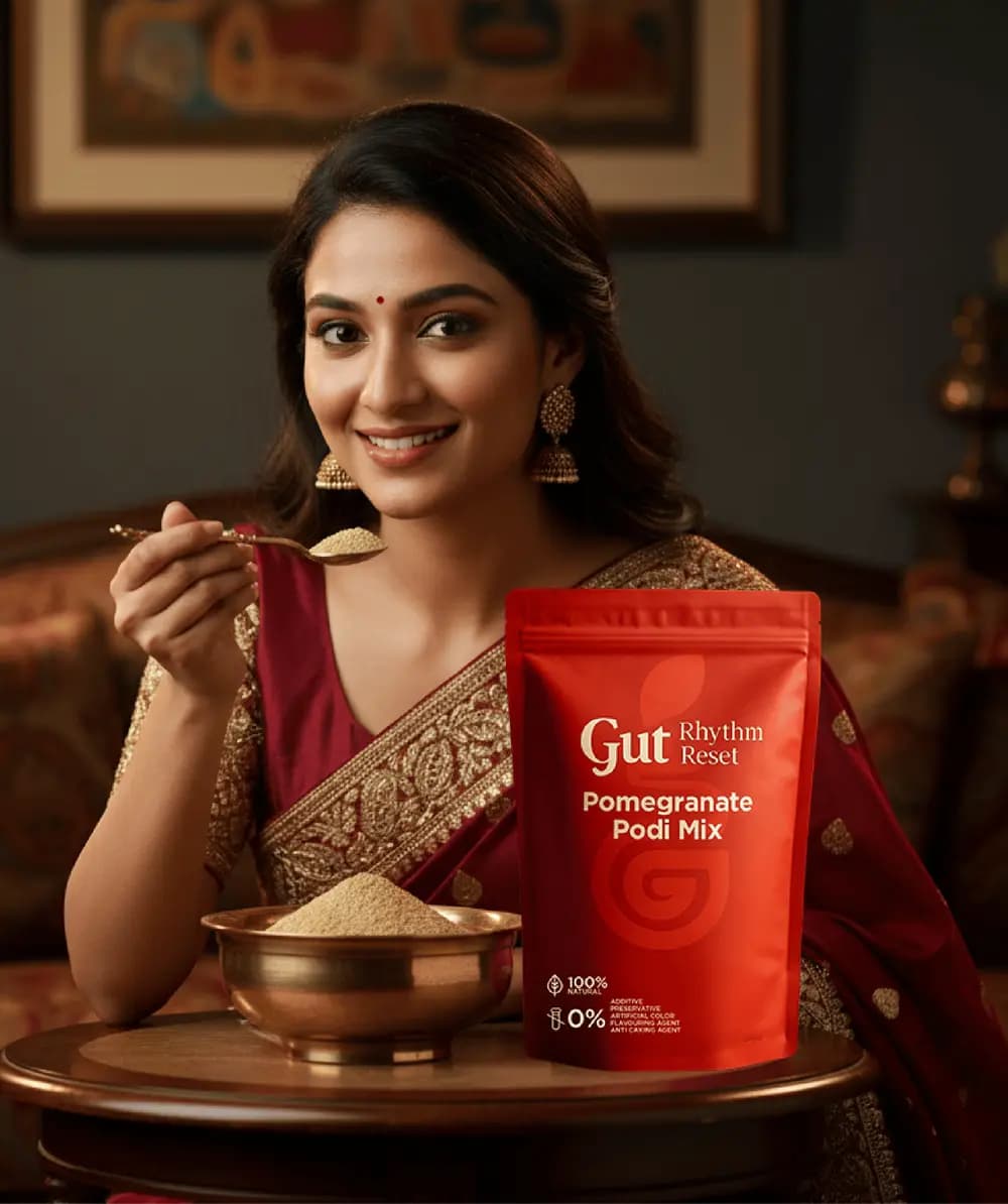 A woman in a red saree with a spoon of Pomegranate Podi Mix, the pouch and a brass bowl on the table